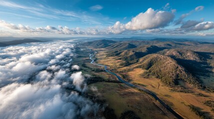 Overhead view captures the breathtaking river valley winding through lush mountains and billowing clouds under a clear blue sky, showcasing nature's beauty and tranquility