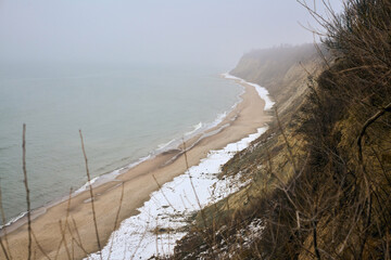 Overlooking misty coastal cliffs with snow-dusted sand and calm sea, creating tranquil atmosphere....