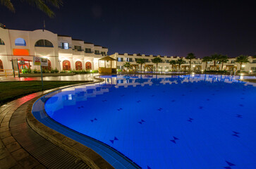 Expansive resort pool glows blue under serene night sky. Lit white buildings surround area, enhancing tranquil atmosphere. Perfect for relaxation and reflection