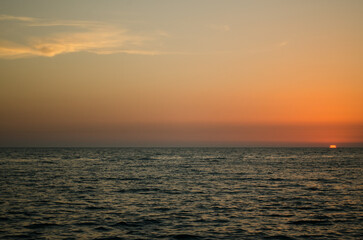 Fototapeta premium Calm ocean scene under warm, gradient sunset sky. Sun partially dips below horizon, casting orange glow. Peaceful atmosphere with gentle waves and expansive view