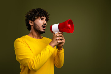 Young man in casual yellow shirt holding a megaphone and expressing surprise against a green studio background