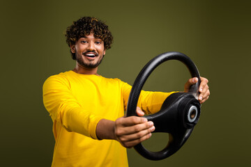 Cheerful young man in a yellow shirt holding a steering wheel with enthusiasm against a khaki green...