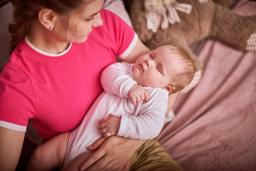 Young woman with braided hair gently cradles sleeping baby girl in pink and beige room. Warm lighting and soft textures create calm atmosphere