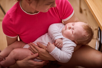 Young woman with blonde hair cradles infant in pink room. Baby girl sleeps peacefully in woman lap. Gentle lighting and warm ambiance enhance soothing scene