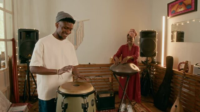 Happy young African American man in casual attire beating percussion and looking at his girlfriend playing glucophone during musical repetition in home studio