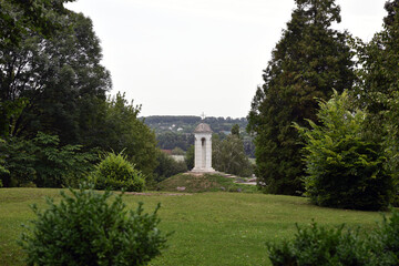 a small bell tower in the park. white bell tower surrounded by green trees. religious premises, Christianity or Catholicism. stone walls, cross on the dome. spring or summer time