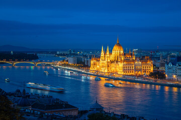 Obraz premium A beautiful night view of the Hungarian Parliament with boats on the Danube River, showcasing Budapest's vibrant lights and reflections