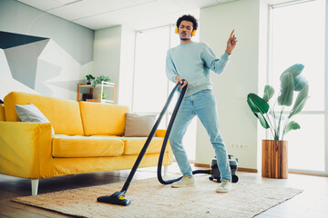 Young man enjoying leisure time vacuuming and moving to music in a stylish living room with bright...