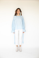 Short hair brunette woman wear blue blouse with puff sleeves and white denim jeans pants, long silver beads earrings, look elegant, casual and smiling. She look at side standing on white background.