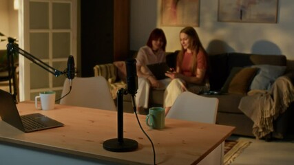 Wooden table with professional equipment and laptop, two talented teenagers sitting on comfortable sofa and discussing new topic of podcast - Powered by Adobe