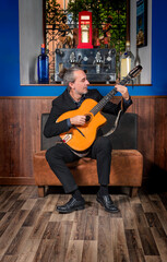 Young jazz musician sitting on an armchair playing an acoustic guitar leaning on his leg, wearing a retro style black suit