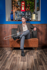 Young jazz musician sitting on an armchair with a trombone resting on his leg, wearing a retro style