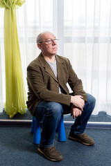 A middle-aged man in a green jacket sits on a chair near a panoramic window. Vertical photo.