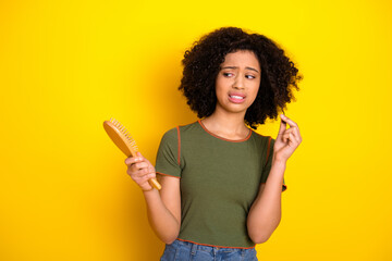 Confused young woman holding a hairbrush against a yellow background, showing dissatisfaction with...
