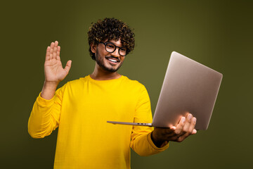 Young man in a yellow shirt waving while using a laptop, smiling, against a khaki background,...