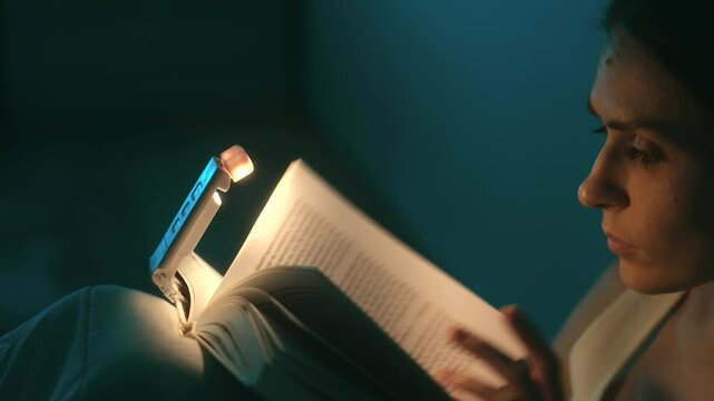 Young woman comfortably reading a paperback book in the dark, using a small portable led clip on light attached to the top of the book, creating a cozy and focused atmosphere