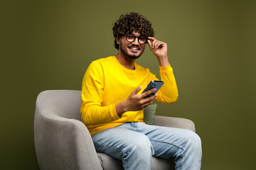 Young man in casual yellow shirt sitting stylishly, holding a smartphone, and smiling confidently...