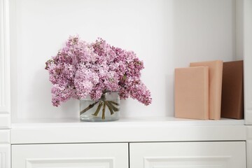 Beautiful lilac flowers in glass vase and books on shelf indoors