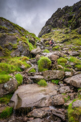 A determined hiker steadily ascends a challenging, rugged rocky Carrauntoohil mountain path that is surrounded by lush greenery and towering cliffs under a moody, dramatic sky overhead, Kerry, Ireland