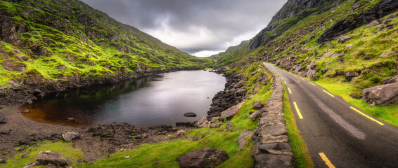 A stunning view showcases a winding road that gracefully hugs the edge of a tranquil lake, beautifully nestled between lush green hills of Gap of Dunloe, all under a captivating sky, Kerry, Ireland