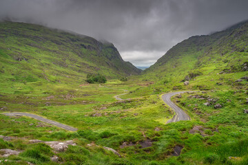 A serene and picturesque green valley showcases a winding road, embraced by majestic hills of Gap of Dunloe and an overcast sky, perfectly capturing the deep tranquility of nature, Kerry, Ireland