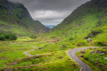 A winding road gracefully cuts through a lush and vibrant Gap of Dunloe valley, flanked by dramatic, towering mountains beneath a captivating sky, creating truly enchanting scenery, Kerry, Ireland