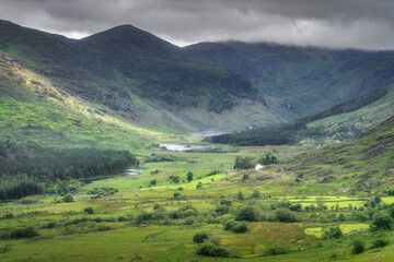A serene and picturesque view of a lush, vibrant Black Valley nestled perfectly between rolling mountains showcases a truly tranquil landscape beneath moody, overcast skies, Kerry, Ireland