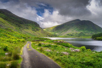 A scenic path winds through lush greenery to a serene lake framed by majestic mountains in Black Valley, under a dramatic sky, creating a mesmerizing natural landscape, Kerry, Ireland