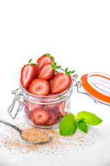 Glass jar with fresh sliced strawberries, brown sugar and mint leaves on white background. Summer dessert ingredients. Clean eating concept. Vertical composition.

