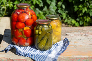 Jars of farm canned cucumbers and tomatoes on wooden table in garden. Pickled homemade vegetables. Copy space. 