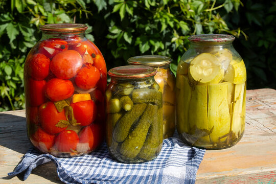 Jars of farm canned cucumbers, zucchini and tomatoes on wooden table in garden. Pickled homemade vegetables. Copy space. 