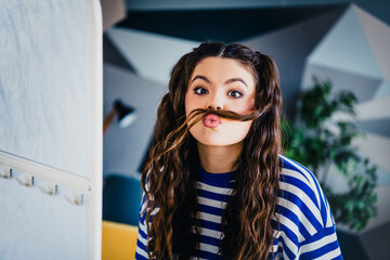 Playful young woman making a mustache from her hair in a casual striped top indoors, expressing a...