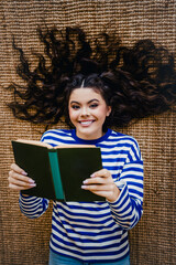 Young woman in striped sweatshirt lying on rug with hair spread and holding book while smiling...