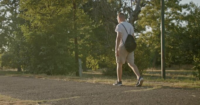 Young man walks and runs on a path through trees in a park during sunset. He carries a drawstring backpack and a water bottle.