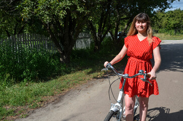 Obraz premium Woman in a red polka dot dress stands with her bicycle on a rural path, surrounded by greenery. The serene atmosphere and natural location reflect outdoor lifestyle and relaxation