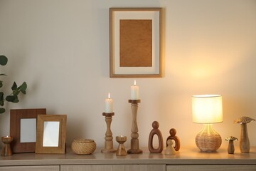 Stylish candlesticks with burning candles and other decor on cabinet near beige wall indoors