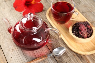 Delicious hibiscus tea in glass cup, teapot, dry roselle sepals and flower on wooden table, closeup