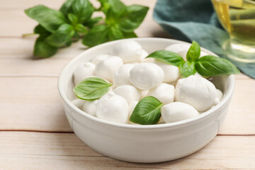 Tasty mozzarella cheese balls in bowl and basil on light wooden table, closeup