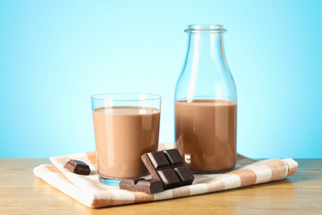 Tasty chocolate milk in glass, bottle and pieces on wooden table against light blue background
