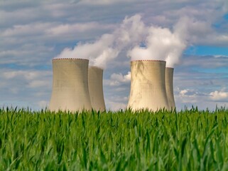 Nuclear power plant cooling towers emitting steam behind a lush green crop field under a partly...