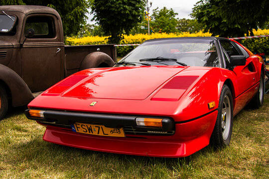Red Ferrari 308 GTS sports car parked on green grass during a vintage car show, with an old rusted truck in the background. Editorial use only. Visible Ferrari logo and license plate.

