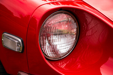 Detailed view of the headlight on a bright red sports car. The chrome and glass reflect light, with...