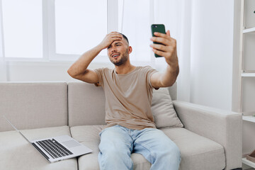Smiling man sitting on a couch, excitedly video calling using a smartphone. Bright, modern living room with a laptop on the side adds to the cheerful atmosphere.