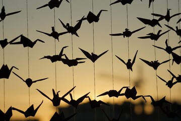 Silhouettes of origami paper cranes hand on strings against a warm, blurred yellow-orange sunset...