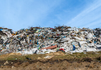 A large mound of garbage is visible at a landfill, consisting of various waste types including plastic bags and discarded furniture, illuminated by sunlight on a clear day