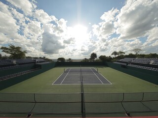 A view of an empty tennis court with green surroundings under a bright cloudy sky on a sunny day