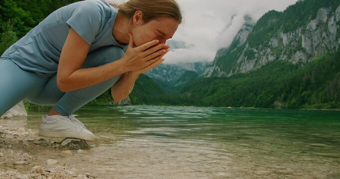 A person drinking fresh water and washing face with clear lake water surrounded by mountains and greenery.