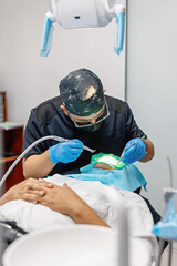 Male dentist working on a patient in a modern dental office. The patient lies relaxed in the chair while the professional conducts a check-up or treatment using gloves and dental tools.