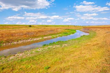 Tranquil waterway meanders through a vast expanse of dry, golden grasslands under a partly cloudy sky. Irrigation ditch reflects the azure sky, a serene path through the dry summer fields