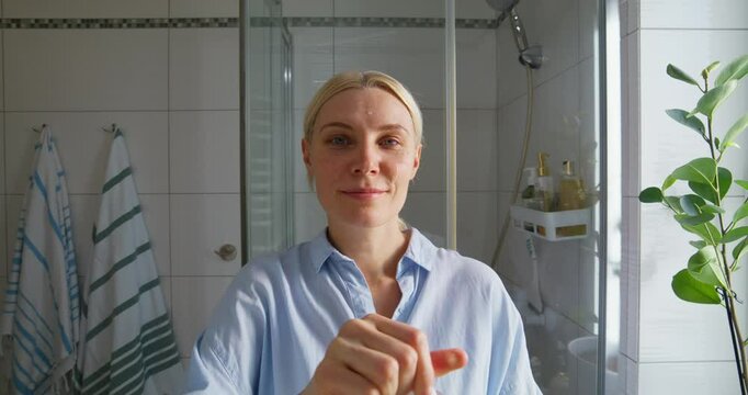 A woman applies a serum or lotion to her face with a pipette. Self-care, morning routine. She looks at the camera standing in the bathroom